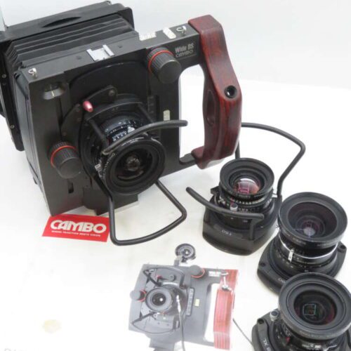 A Cambo Wide DS camera system with a wooden handle, accompanied by three interchangeable lenses placed on a white surface. A brochure titled 'Digital Wide DS System' is visible underneath the camera system, showing an image of the equipment.