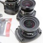Two large camera lenses on a white surface, with one partially over a brochure image of a camera. The lenses are wide-angled, featuring multiple rings and engravings indicating specifications. A coiled cable and ancillary lens equipment are also slightly visible.