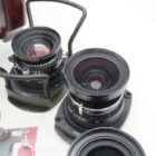 A close-up view of two camera lenses with visible focus and aperture controls, mounted on a white surface. The lenses have black cylindrical bodies with brand markings. Adjacent to the lenses is a section of camera equipment with a wooden handle and an image of a camera partially in view.