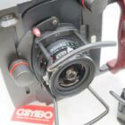 A close-up of a professional Cambo camera featuring a wide-angle lens, with a wooden handle on the right and a detachable lens to the side. The background shows a red Cambo logo card.