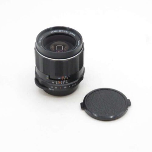A camera lens labeled "ASAHI OPT. CO. JAPAN" with an attached aperture ring and focus indicators, placed on a white surface next to its detached lens cap.