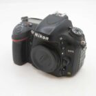 A Nikon D600 DSLR camera body with a lens cap on a white background, featuring various control dials and buttons.