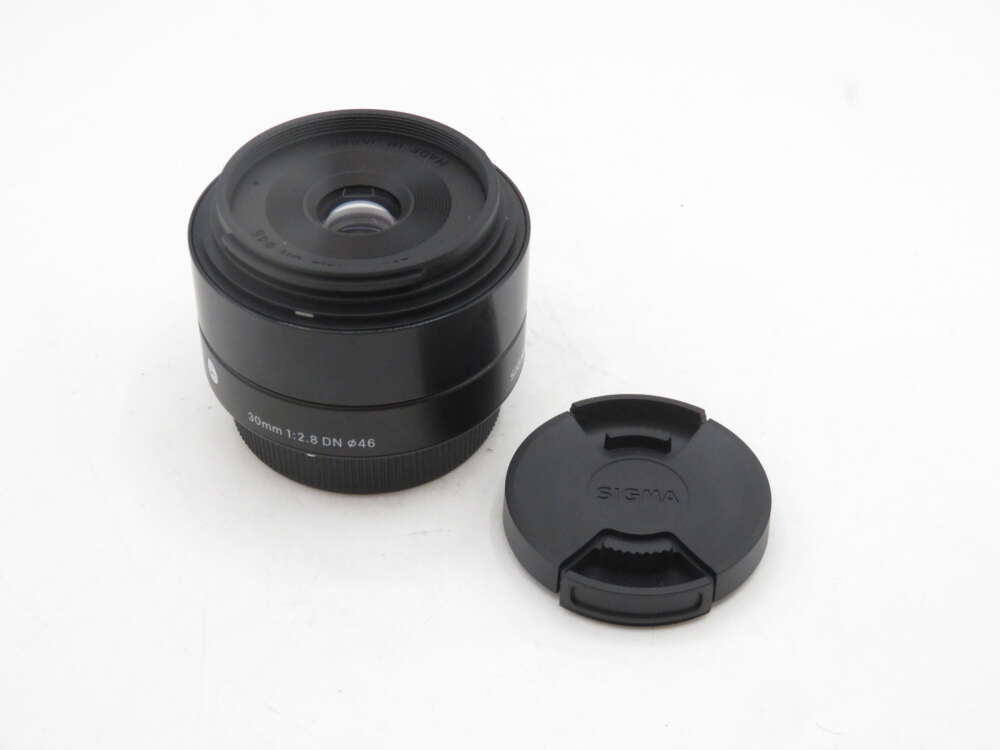 IMG_2099 A Sigma 30mm f/2.8 DN lens placed on a white surface next to its lens cap.