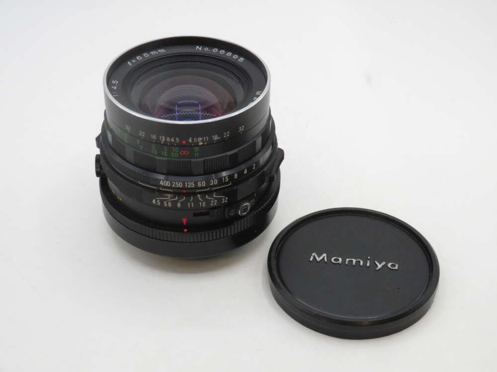 IMG_2146 A Mamiya camera lens with an attached focus and aperture ring, alongside a black lens cap labeled "Mamiya."