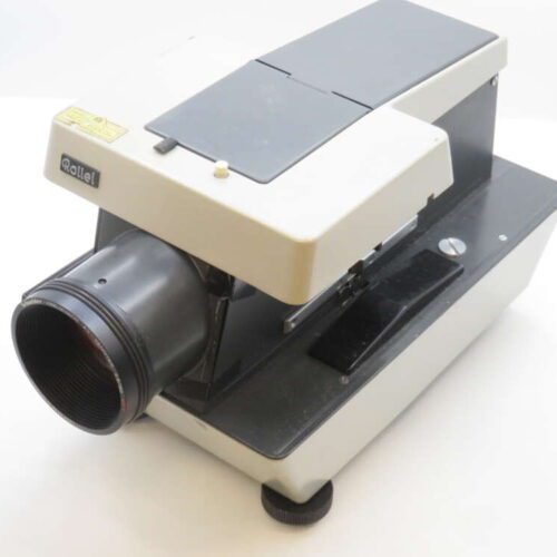 A vintage Rollei slide projector with a black and beige body, featuring a prominent lens at the front and a large, flat top surface. The projector is positioned on a white background, highlighting its retro design and mechanical components.
