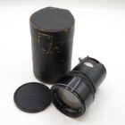 A vintage camera lens with a black metal body, accompanied by a protective carrying case and a lens cap with the logo 'OMEGON'.