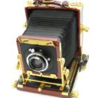 A vintage large format camera with a dark bellows design, polished wood frame, and gold-tone metal fittings, featuring a prominent lens at the front.