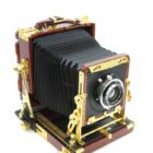 A vintage large format camera with a wooden body, gold metal fittings, and a black bellows extending from the lens.