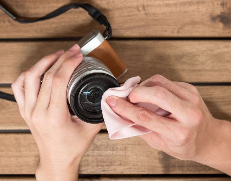 Hands cleaning a camera lens with a pink cloth on a wooden table.