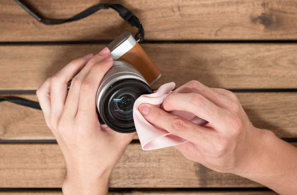 Hands cleaning a camera lens with a pink cloth on a wooden table.