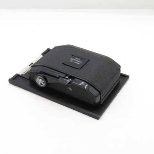 A vintage camera film back with a black textured surface and a dial on the side, placed on a flat surface.