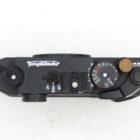 Top view of a black Voigtländer camera, showing dials and controls on a white background.