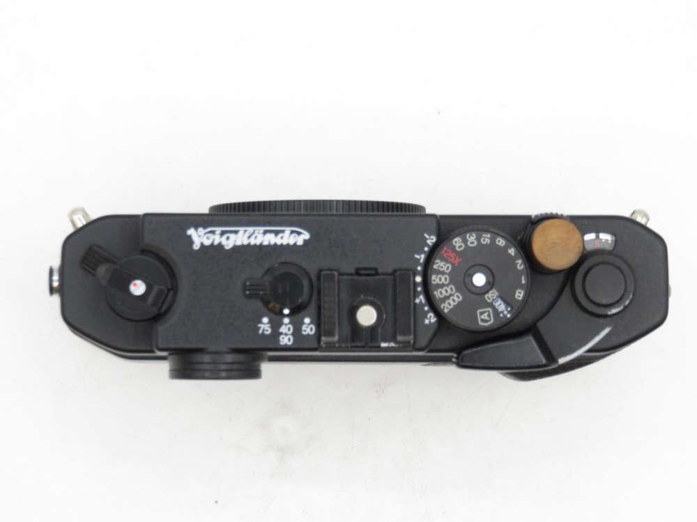 Top view of a black Voigtländer camera, showing dials and controls on a white background.