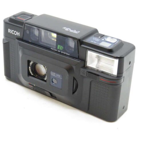 Black Ricoh film camera with a lens cover, visible flash, and branding on the top left.