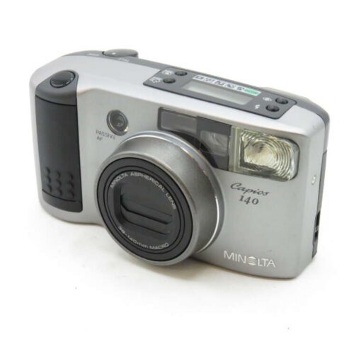 A silver Minolta Capios 140 camera with a zoom lens and built-in flash on a white background.