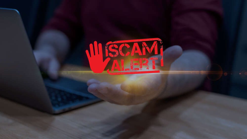 A person sitting at a laptop with a digital "Scam Alert" sign featuring a red hand projected above their hand.