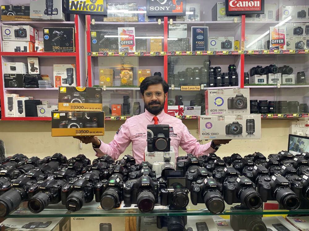 A man stands behind a counter displaying various camera models, holding two camera boxes in a store filled with camera accessories.