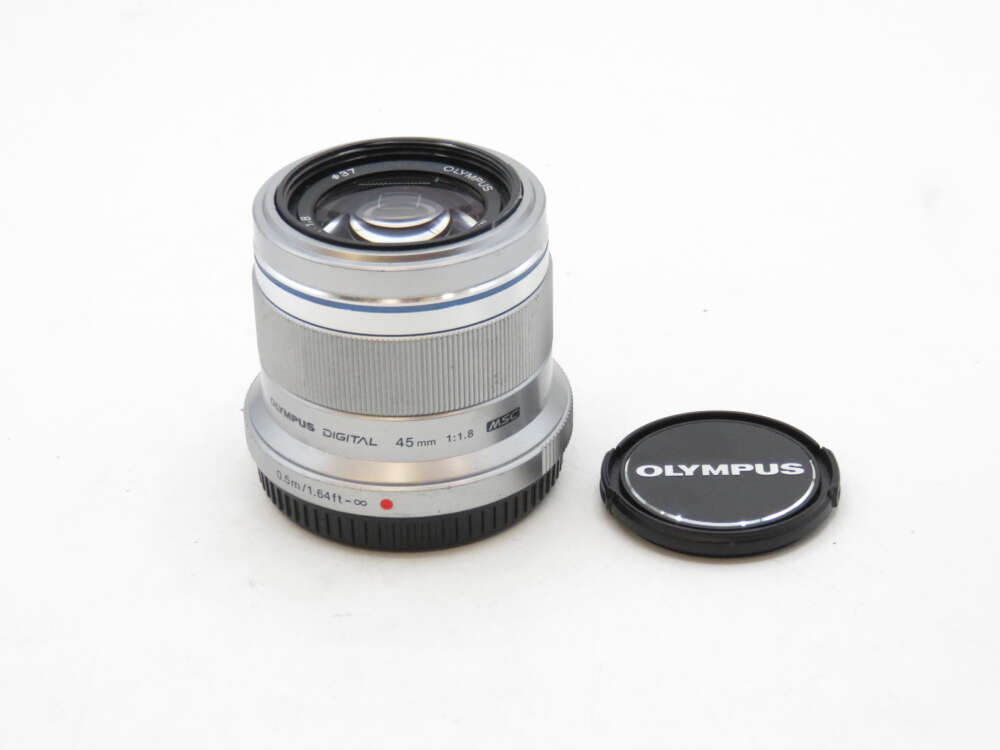 IMG_3412 A silver Olympus 45mm f/1.8 camera lens with its cap resting on the surface beside it.