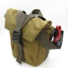 A khaki-coloured canvas bag with a front flap, secured by a brown strap and buckle, and a red carrying handle on the side.