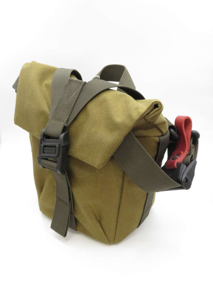 IMG_3738 A khaki-coloured canvas bag with a front flap, secured by a brown strap and buckle, and a red carrying handle on the side.