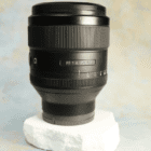 A black camera lens marked as FE 1.4/85 GM, resting on a piece of white styrofoam against a soft, multicoloured background.