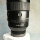 A black camera lens labeled "FE 1.8/135 GM" is placed on a white textured base against a soft, blurred background.