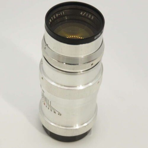 A silver camera lens with an engraved inscription, marked as 4/135, positioned upright on a white background.