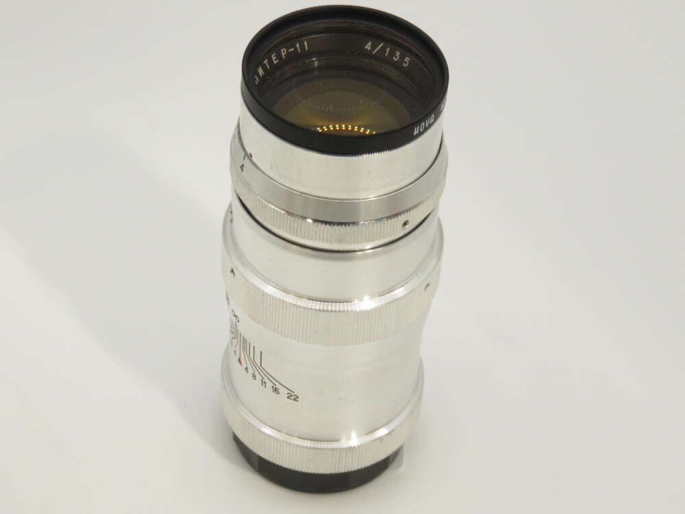 IMG_3820 A silver camera lens with an engraved inscription, marked as 4/135, positioned upright on a white background.