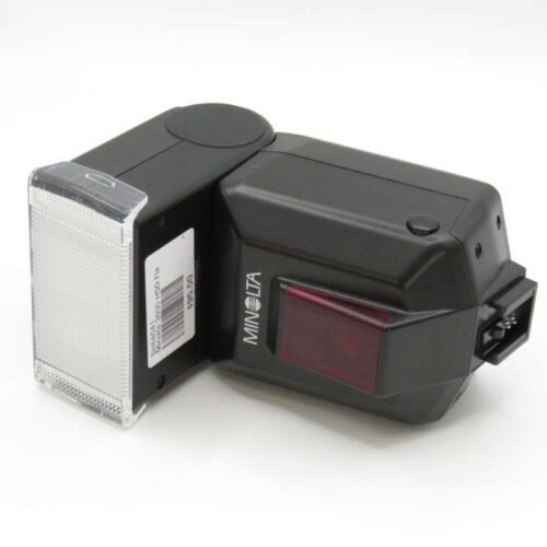 A black Minolta camera flash with a rectangular diffuser and a red sensor panel, placed on a plain surface.