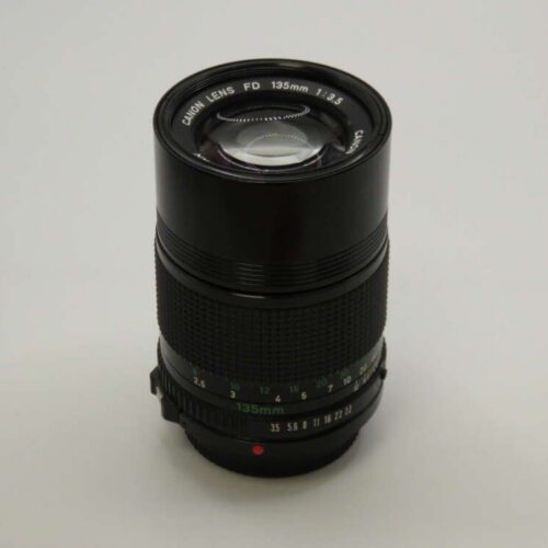 A black Canon FD 135mm f/3.5 camera lens with focus and aperture rings, viewed from above on a plain background.