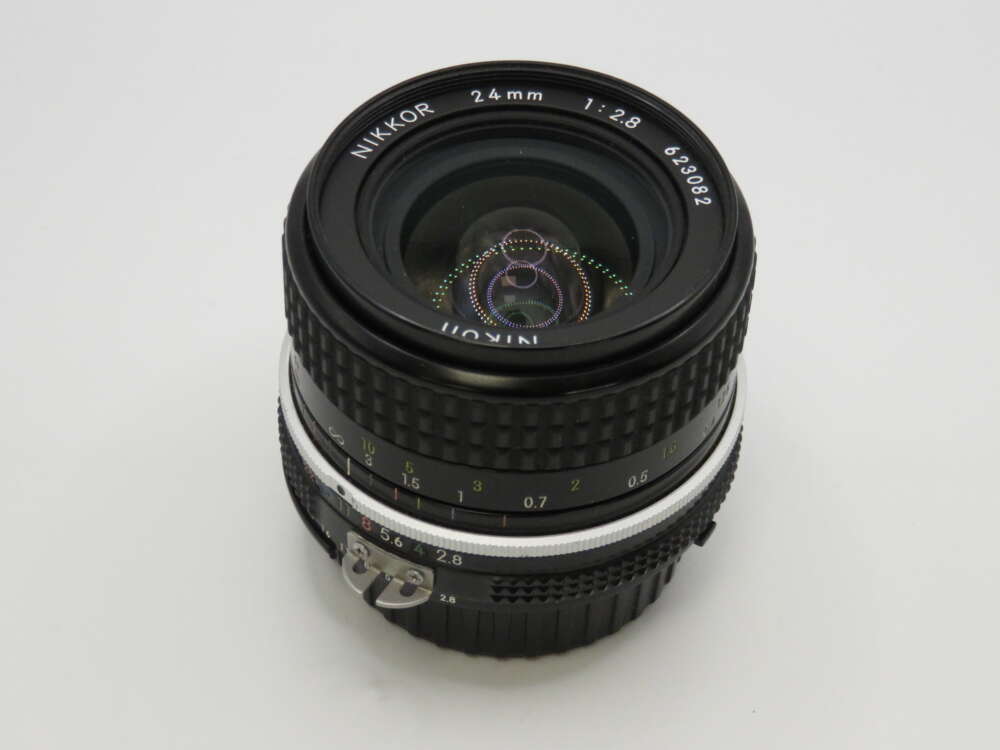 IMG_4015 A Nikon Nikkor 24mm f/2.8 camera lens with a black ribbed exterior and clear glass elements reflecting light.