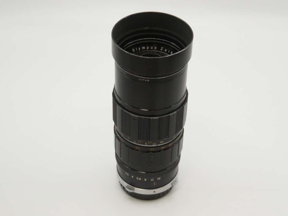 IMG_4024 A dark Olympus zoom camera lens with multiple adjustment rings and focal length markings on a plain background.