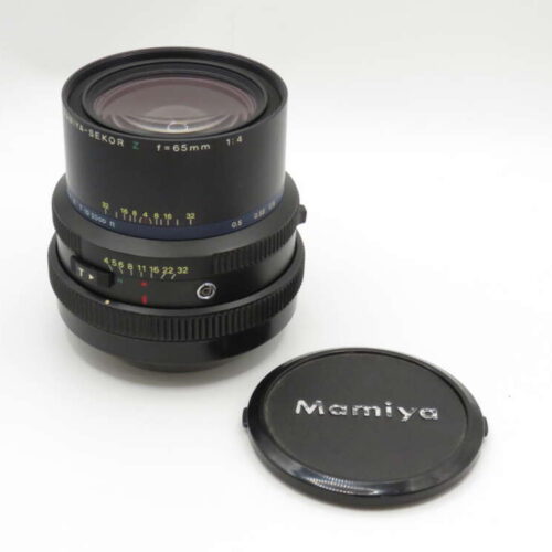 A Mamiya Sekor Z 65mm f/4 lens sits upright beside a Mamiya lens cap on a plain surface.