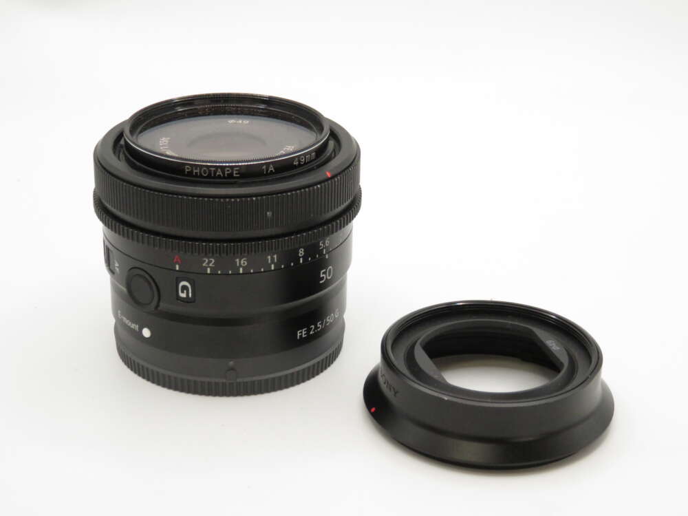 IMG_4199 A camera lens with an attached hood, both black, resting on a white surface.