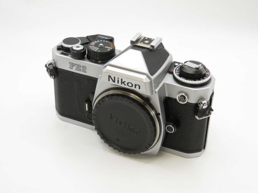 IMG_4221 Silver and black Nikon FE2 film camera with a Vivitar lens cap, placed on a plain white background.