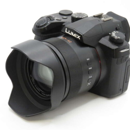 A black Lumix camera with a wide lens and a lens hood, viewed from an angle, on a white background.