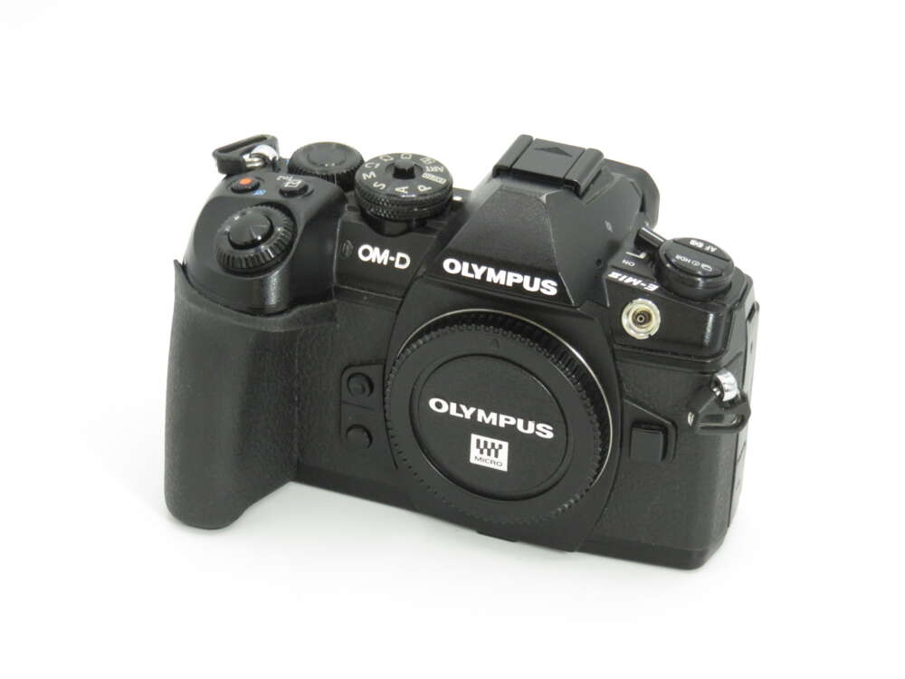 IMG_4323 A black Olympus OM-D digital camera with various control dials and buttons, placed on a white surface.